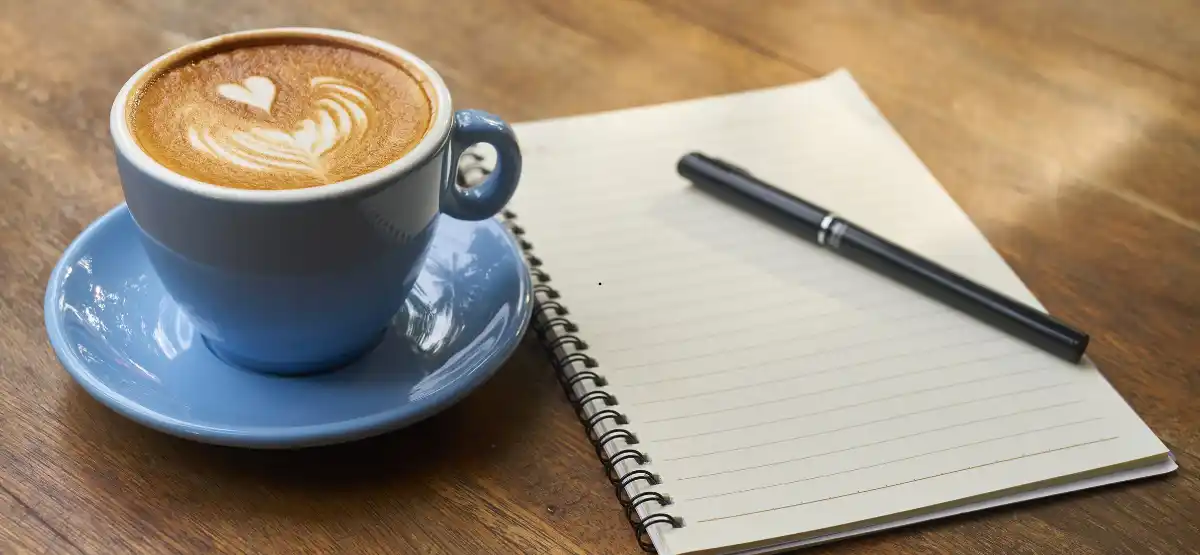 A blue cup of cappuccino with heart-shaped latte art sits on a wooden table next to an open, blank spiral notebook and a black pen.