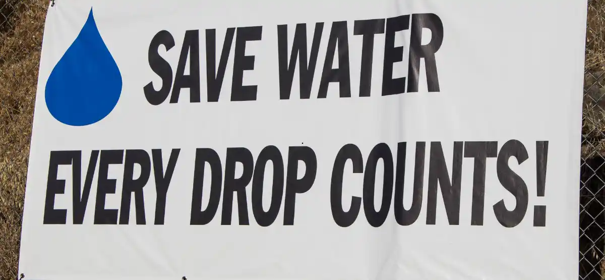 A banner with a large blue water droplet icon and bold text that reads: "SAVE WATER EVERY DROP COUNTS!" displayed outdoors.