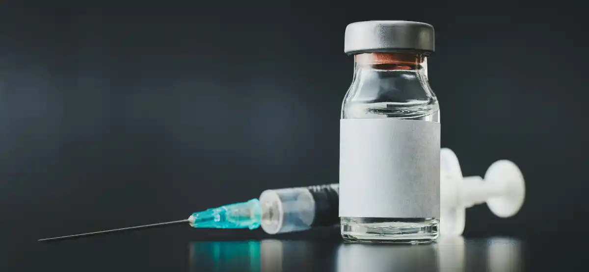 A small glass vial with a blank white label stands upright next to a syringe with a needle, set against a dark background.