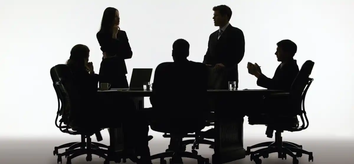 Silhouettes of five people in business attire sitting and standing around a conference table, engaged in discussion in a meeting room with a white background.