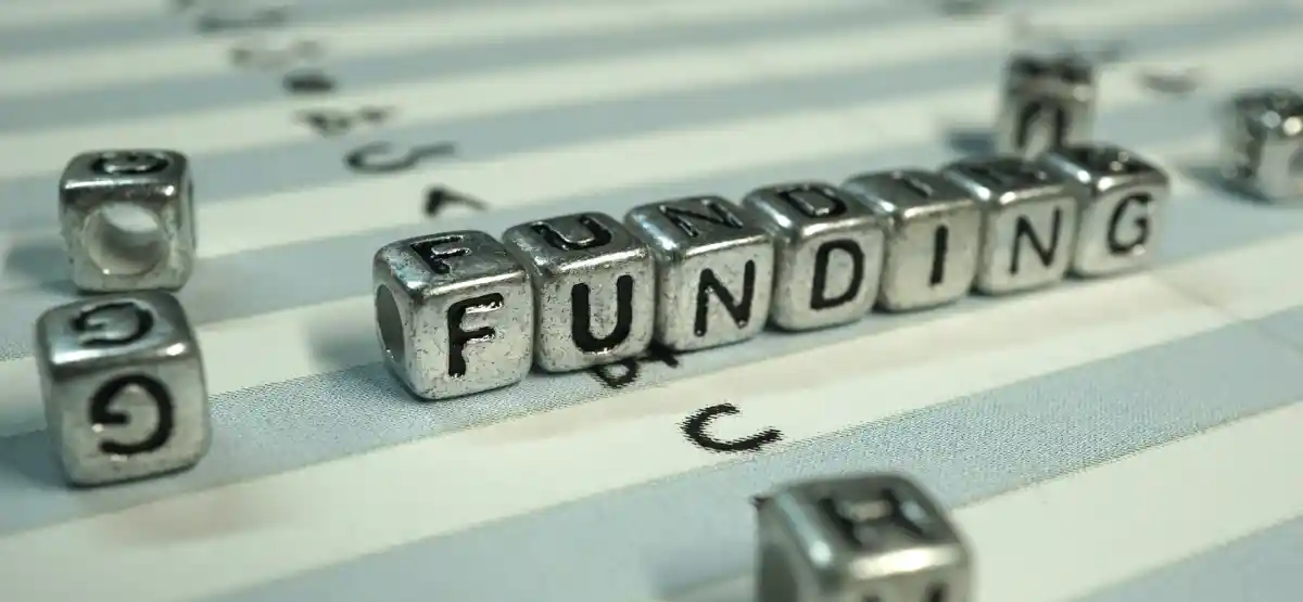 Metallic letter blocks spell out "FUNDING" on a striped, light-colored surface, with several other scattered blocks and letters visible in the background.