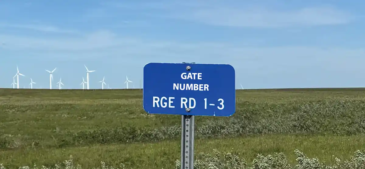 A blue sign reads "Gate Number RGE RD 1-3" in white text, standing in a grassy field with wind turbines and a blue sky in the background.